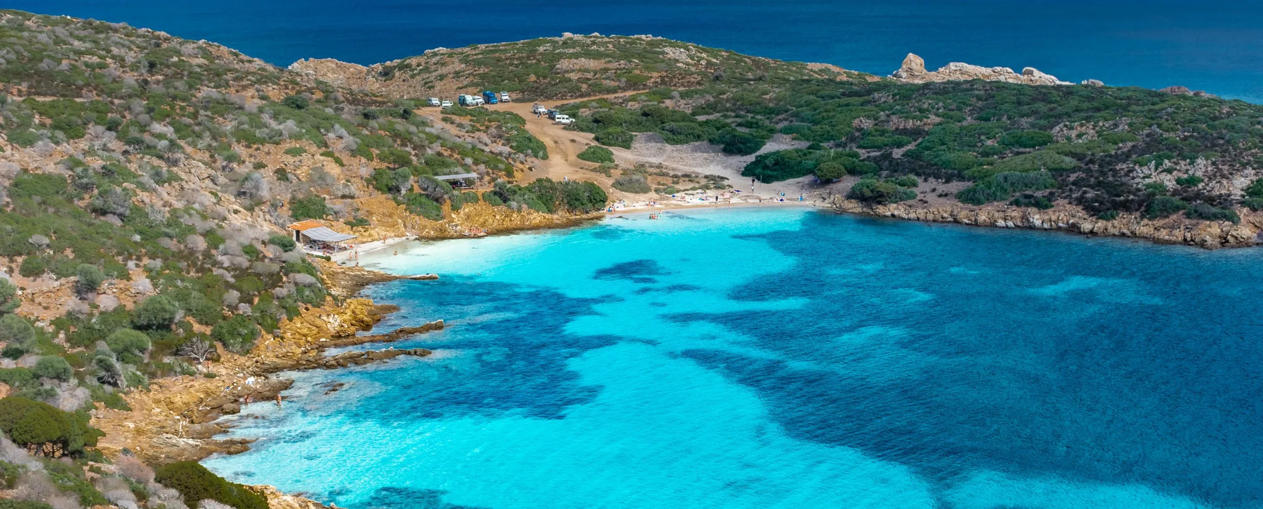 The image beautifully captures a serene bay with strikingly clear turquoise water contrasting with the deeper blue of the open sea. The sandy beach is visible, backed by a scrubland hill. The overall impression is one of natural beauty and tranquility, perfect for a travel or nature theme.