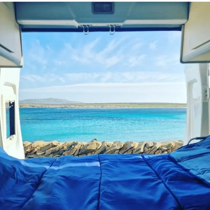 The image captures a beautiful and serene view of a blue ocean and sky from the perspective of inside a van, with a comfortable blue blanket in the foreground. The composition effectively frames the expansive landscape.