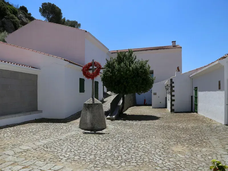 An outdoor shot of a white-washed building complex featuring a cobblestone courtyard. A prominent stone cross adorned with a red wreath stands in the foreground, next to a small tree casting shadows. The scene is set under a clear blue sky.
