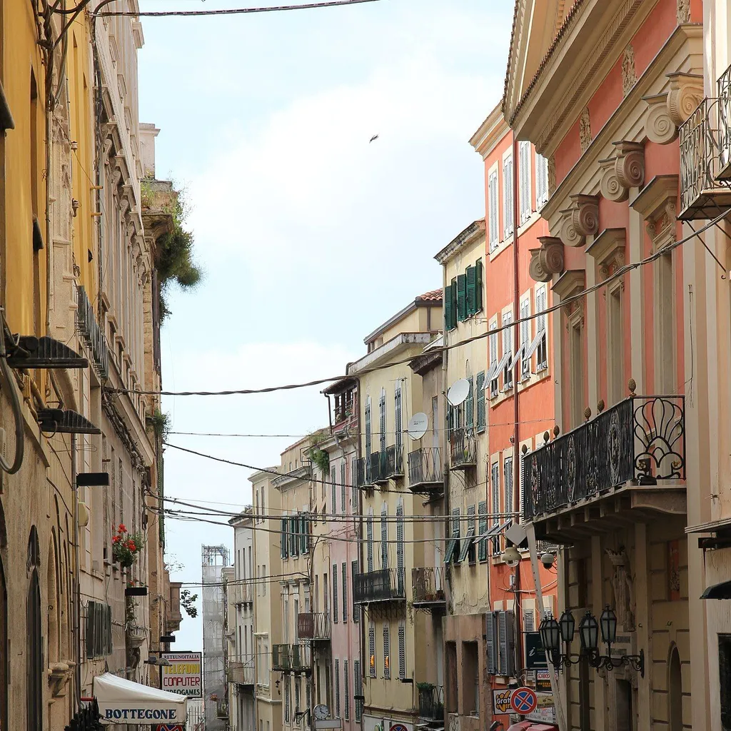 An editorial image showcasing a charming, narrow street lined with colorful, traditional buildings. The street slopes gently downhill, creating depth, and features various architectural details, shop signs, and some pedestrians. Overhead wires are prominent throughout the scene.