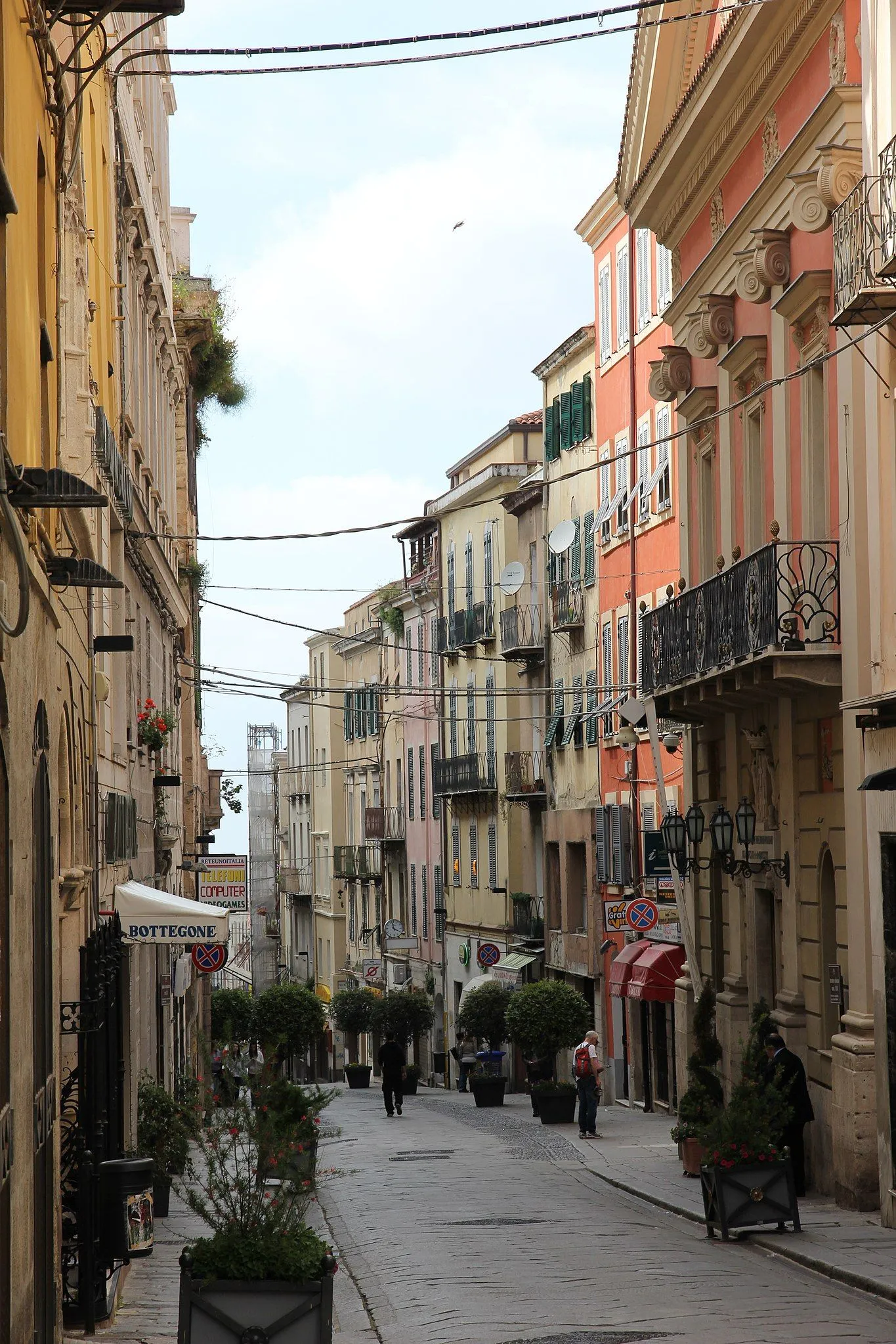 An editorial image showcasing a charming, narrow street lined with colorful, traditional buildings. The street slopes gently downhill, creating depth, and features various architectural details, shop signs, and some pedestrians. Overhead wires are prominent throughout the scene.