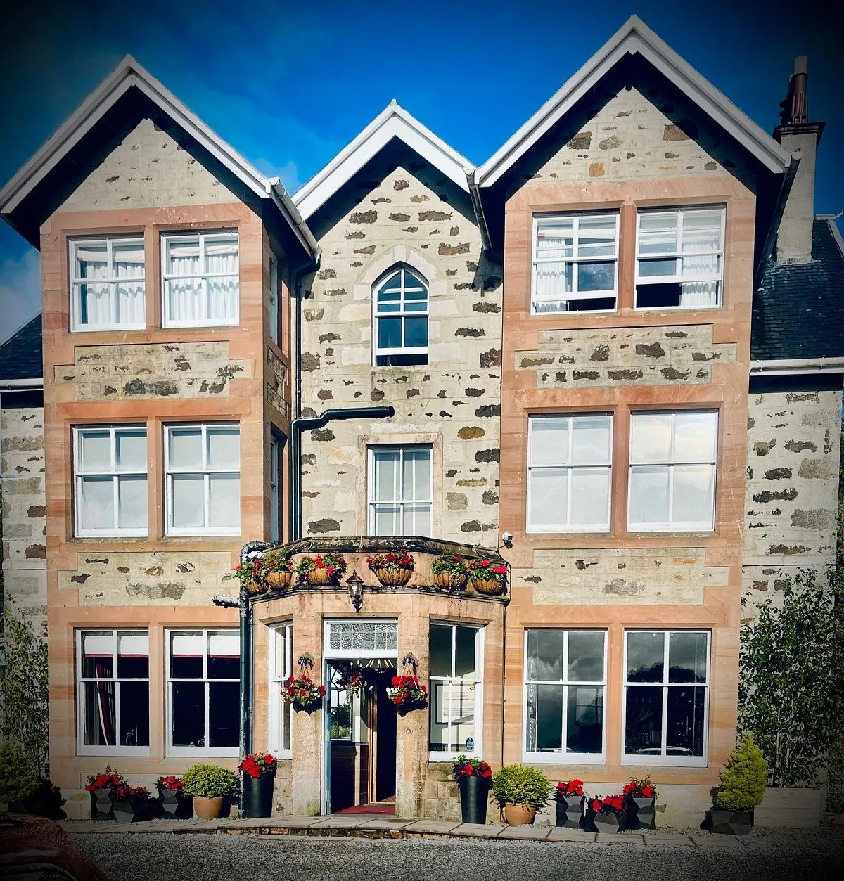 An editorial quality image depicting the stone facade of a building with multiple windows and an inviting entrance adorned with hanging flower baskets and potted plants. The sky above is a vibrant blue.