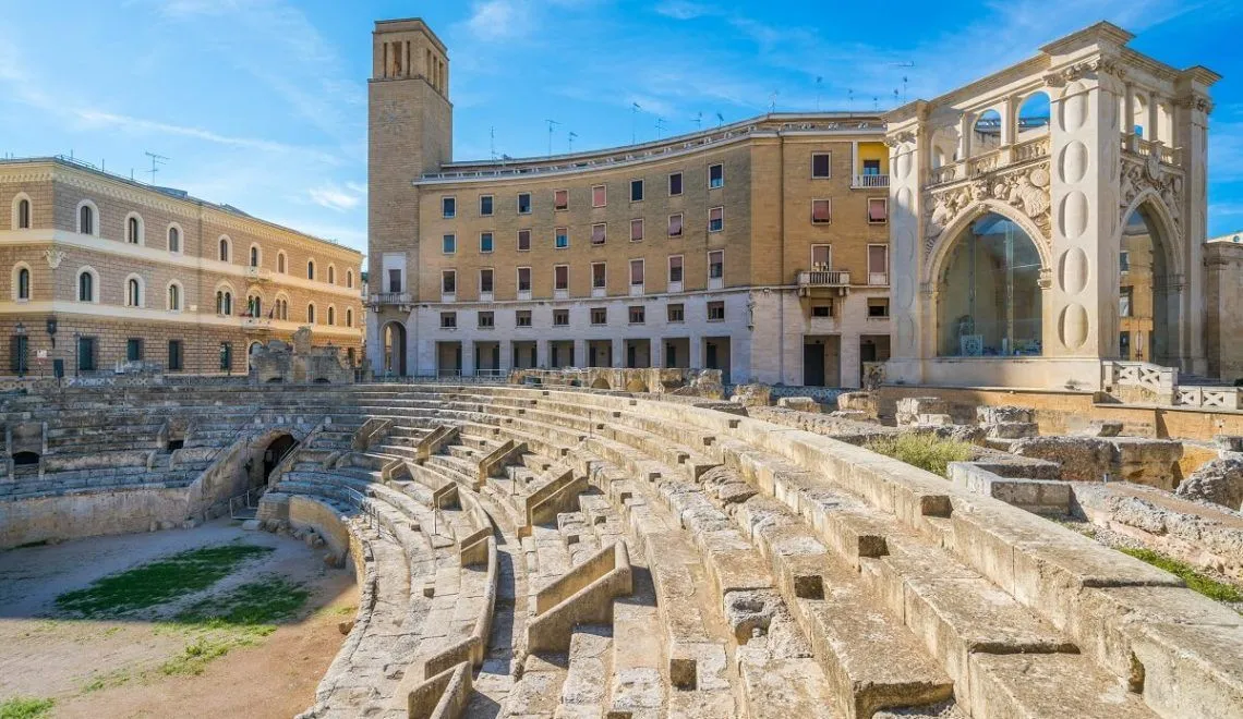 An editorial style image showcasing the ancient Roman amphitheater ruins with modern and historic buildings in the background under a clear blue sky.