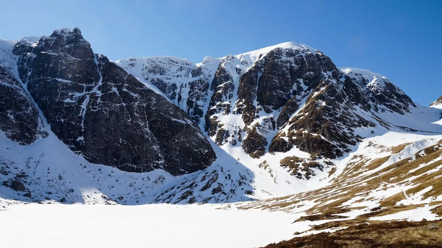 A majestic and vast snow-covered mountain range stands tall under a clear, brilliant blue sky. The rugged rock faces are partially covered in pristine white snow, with deep shadows and highlights defining the terrain's contours. The foreground is a wide expanse of untouched snow, leading up to the imposing peaks.