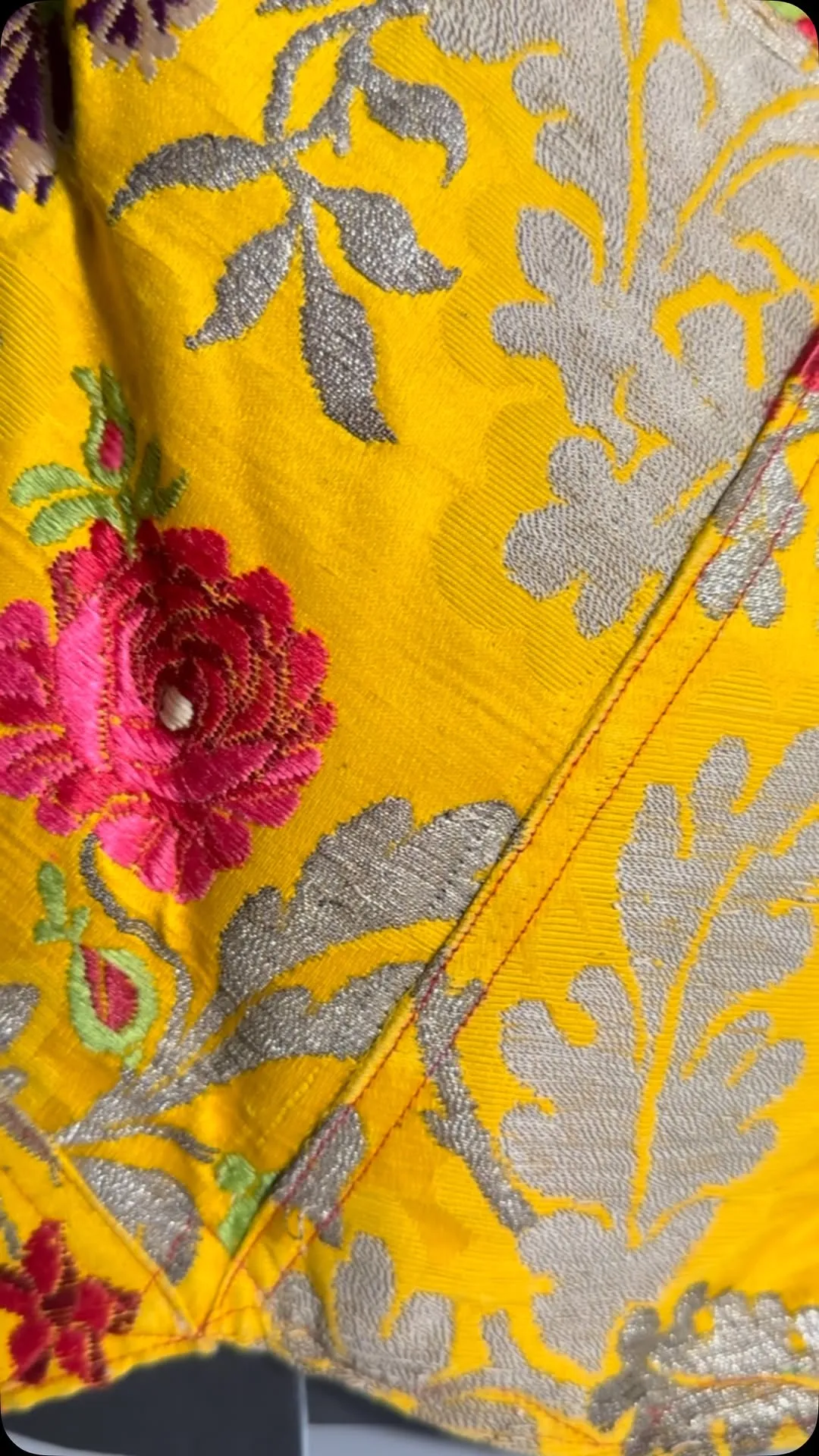 A vibrant close-up shot of yellow fabric adorned with intricate pink floral embroidery and subtle grey metallic patterns. The texture of the fabric and the details of the embroidery are clearly visible.
