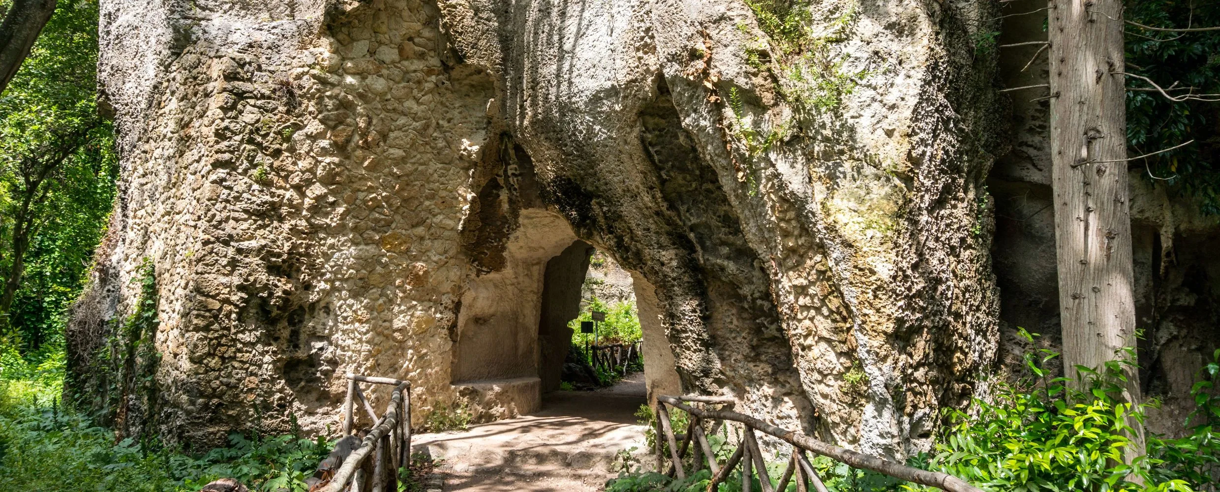 A view through a stone archway or grotto, revealing a sun-dappled path lined with wooden railings and green foliage. The natural rock formations and an opening to another area create a sense of discovery and depth.