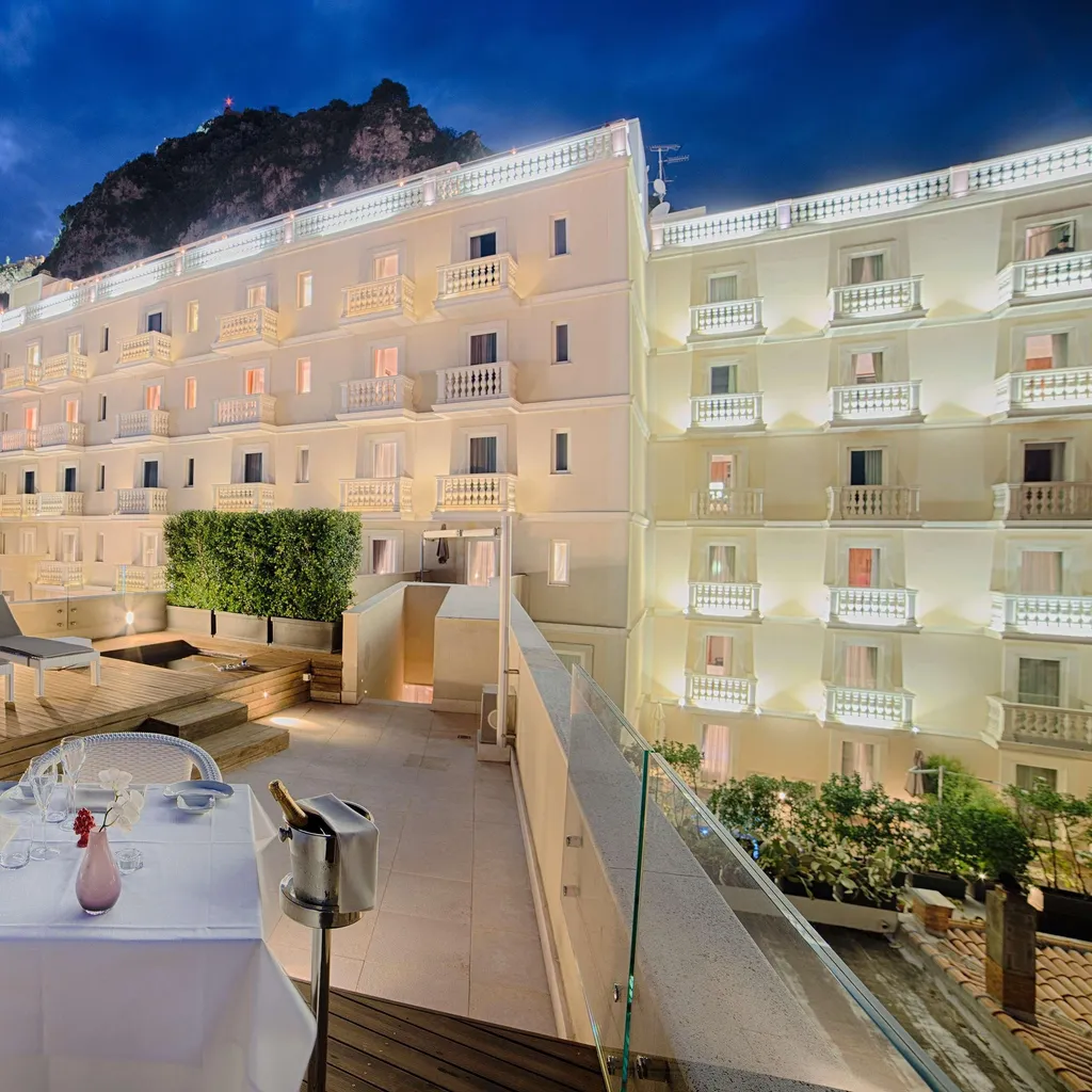The image displays a well-composed and highly photo-realistic hotel rooftop terrace at dusk, featuring a dining table setup in the foreground and sun loungers. The hotel building is brightly lit against the dark blue sky and a mountain backdrop.