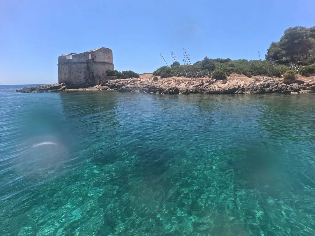 This image showcases a beautiful coastal scene with clear turquoise water, a rocky island, and an old, rustic building under a bright blue sky. The vibrant colors of the water and sky are particularly striking, and the composition effectively captures the serene atmosphere of the location.