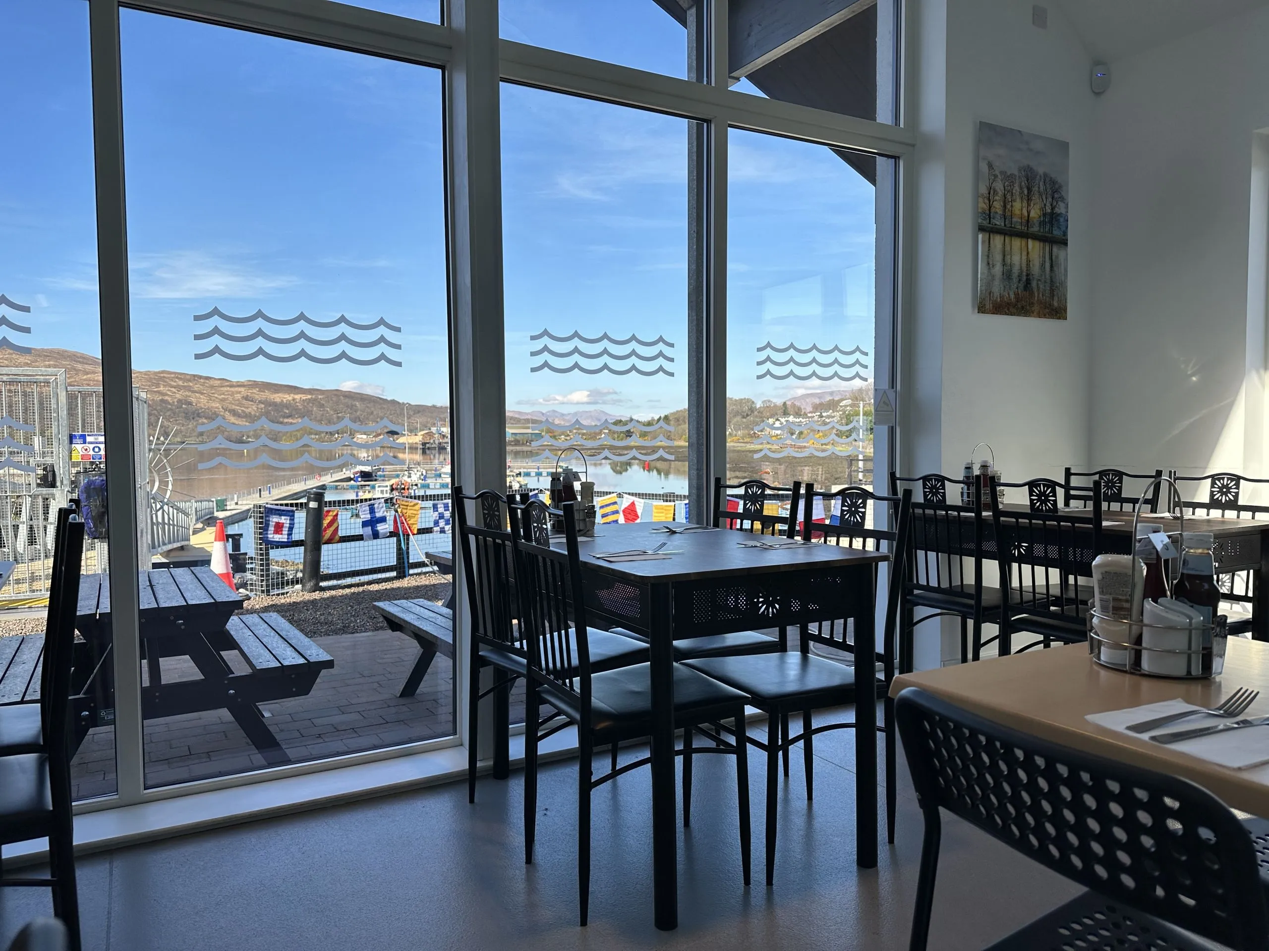 An interior shot of a cafe or restaurant featuring large windows with a view of a body of water, distant hills, and a dock. Several dining tables and chairs are visible in the foreground and midground.