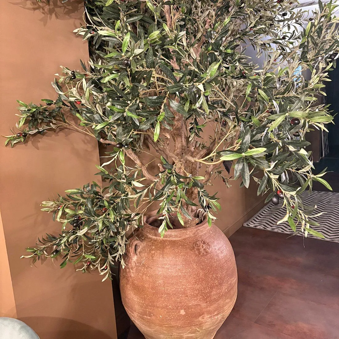 The image features a large artificial olive tree in a rustic pot, placed indoors against a brown wall. The composition is focused on the plant, which occupies most of the frame. The lighting is even and natural, highlighting the details of the leaves and the texture of the pot. The colors appear accurate, and the image is generally sharp with the key elements in focus, contributing to its photo-realistic quality.