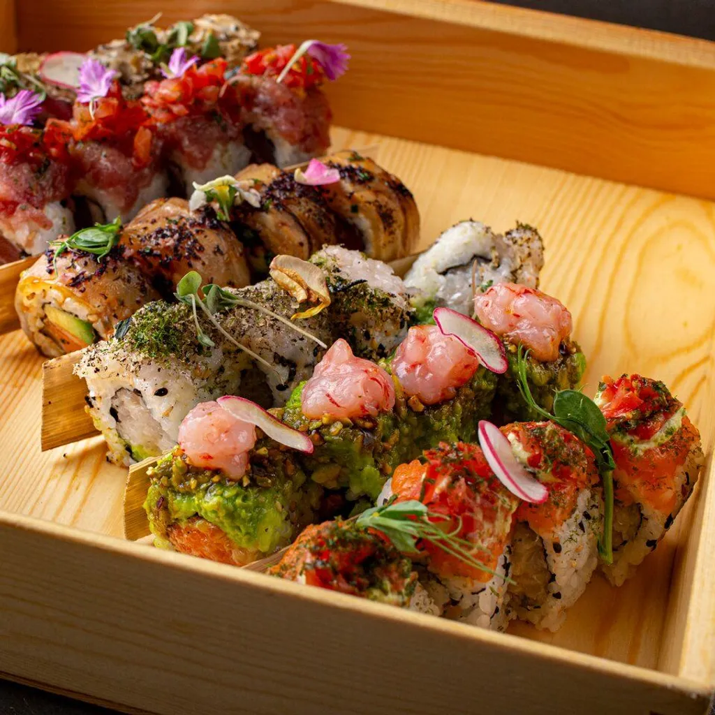 An editorial-style image showcasing a variety of intricately prepared sushi rolls presented in a rustic wooden tray. The rolls feature diverse toppings and garnishes, including various types of fish, avocado, herbs, and edible flowers, arranged artfully to highlight their details and textures.
