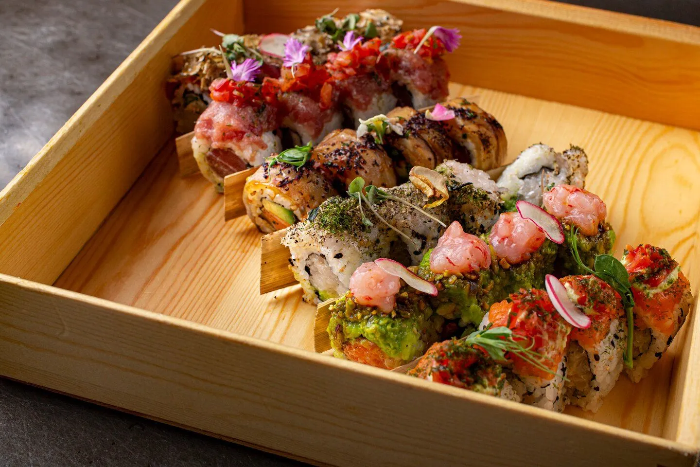 An editorial-style image showcasing a variety of intricately prepared sushi rolls presented in a rustic wooden tray. The rolls feature diverse toppings and garnishes, including various types of fish, avocado, herbs, and edible flowers, arranged artfully to highlight their details and textures.