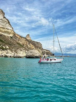 A white sailboat with people on board is anchored in clear turquoise water beside a large, light-colored rocky cliff under a blue sky with scattered clouds.