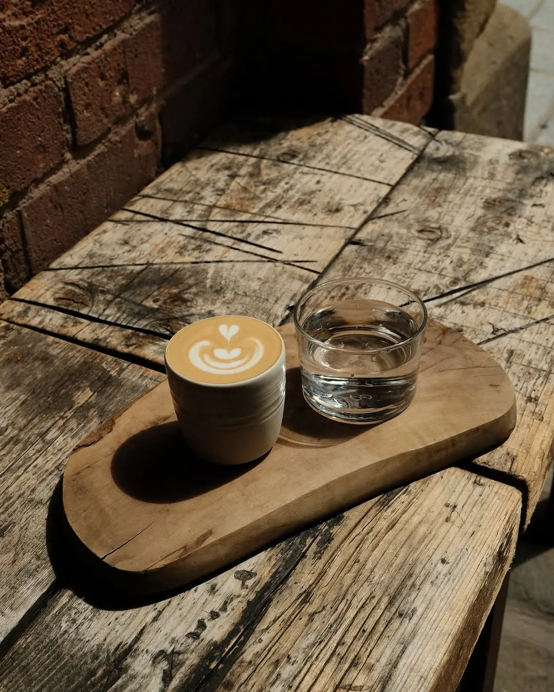 An inviting overhead shot of a latte with delicate latte art and a glass of water, presented on a rustic wooden board. The natural lighting casts interesting shadows, highlighting the textures of the wood.
