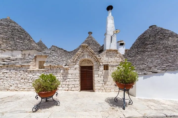 A picturesque view of traditional trulli houses in Puglia, Italy, featuring their distinctive conical stone roofs and white-washed walls under a clear blue sky, with potted plants in the foreground.