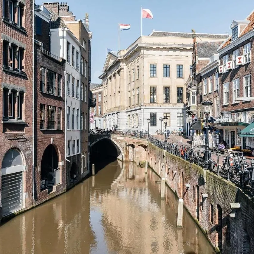 An inviting canal scene in a European city, featuring historical architecture, bridges, and clear reflections on the water, captured under bright daylight.