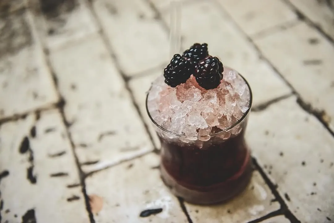 A close-up image of a vibrant berry-based cocktail or iced drink, generously topped with crushed ice and fresh, dark blackberries. The drink is served in a clear glass and set against a light-colored, textured tiled surface, creating an inviting and refreshing visual.