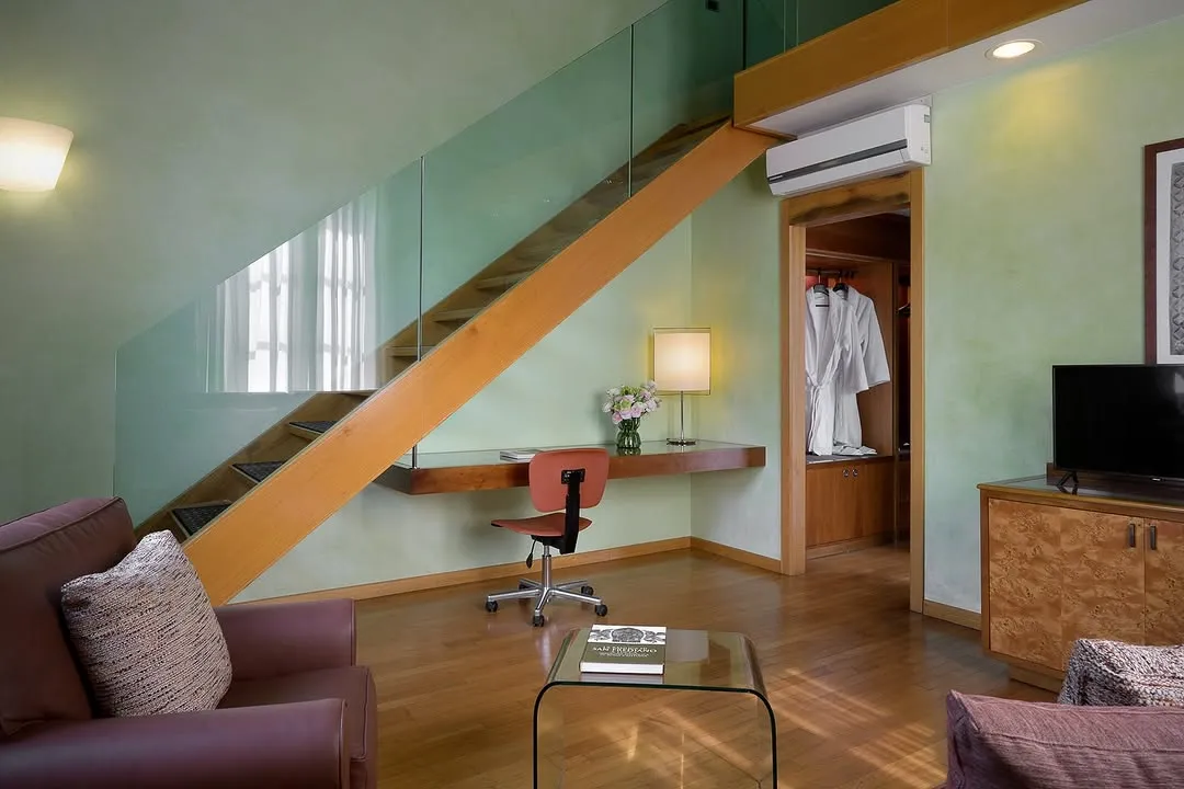 A well-composed and sharply focused image of a modern interior space, likely a hotel room or apartment, featuring a wooden staircase with a glass railing, a study desk area with a chair, and part of a living area. The lighting is good, and colors appear natural and accurate.