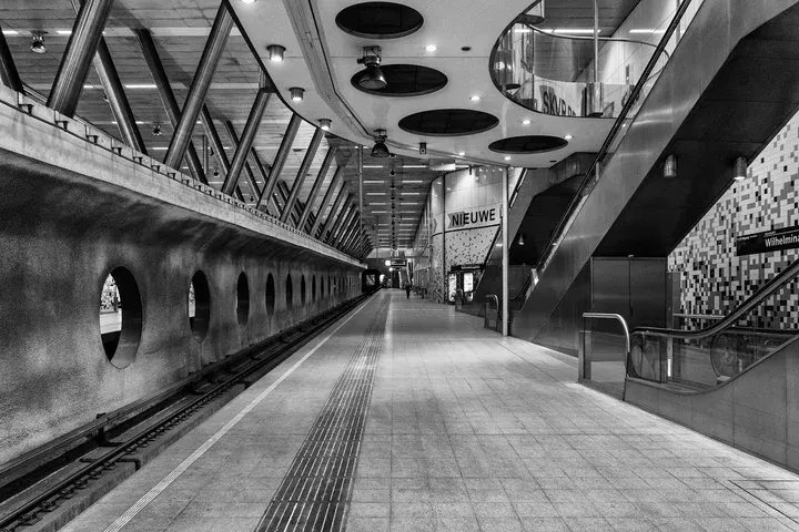 A well-composed, sharp, and photo-realistic black and white photograph of a modern subway station. The image effectively uses leading lines and architectural elements to convey depth and structure. The lighting is atmospheric and appropriate for the underground setting, creating strong contrasts. The tonal range is excellent, and all key elements from foreground to background are in sharp focus.