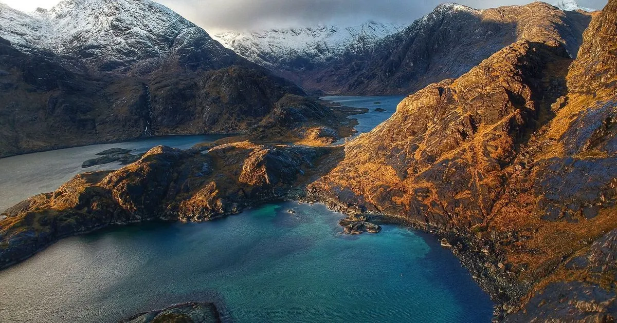 An aerial shot showcasing a breathtaking natural landscape with majestic snow-capped mountains and a winding blue-turquoise lake or fjord. The scene is illuminated by dramatic natural light, casting long shadows and highlighting the rugged textures of the terrain.