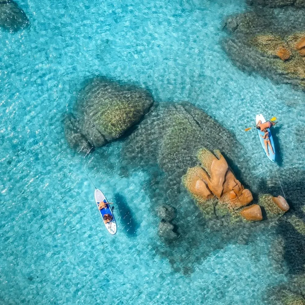 An aerial view captures two individuals enjoying paddleboarding in crystal-clear turquoise waters amidst large, natural rock formations.