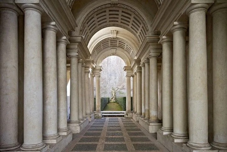 An image capturing a long, vaulted hallway lined with classical columns, leading to an arched opening revealing a statue in a garden area beyond. The floor features a patterned mosaic.