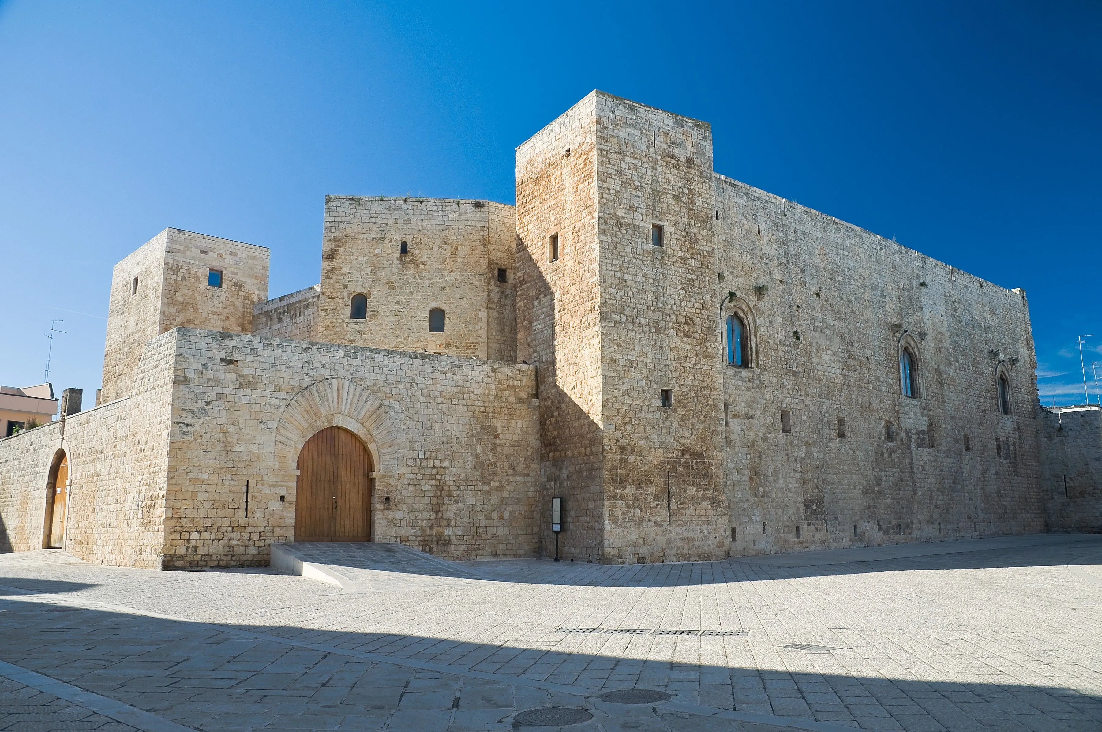 An exterior view of a large, ancient stone castle or fortress under a clear blue sky. The image highlights the sturdy stone walls, a prominent arched entrance, and a paved courtyard.