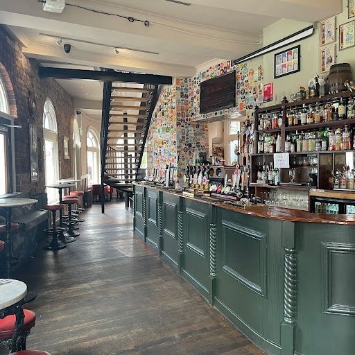 This image showcases the interior of a pub, specifically the bar area. The composition is well-balanced, leading the viewer's eye towards the bar counter. The lighting is good, providing a well-lit and inviting atmosphere. The image is sharp, capturing details of the wooden floor, brick walls, and the bar's intricate design. Color accuracy is good, with natural-looking tones and a pleasant color palette. The bar counter is the key element in focus, and it is presented in a photo-realistic manner. The image is free from any unwanted elements, such as clutter, text, or promotional graphics. Overall, the image is well-composed, well-lit, sharp, and color-accurate, making it a high-quality representation of the pub's interior.