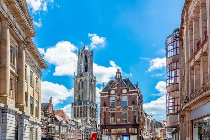 A well-lit and sharp photograph showcasing the architecture of a European city. The main tower is a clear focal point, framed by other historic buildings against a vibrant blue sky with clouds. Colors are accurate and the image is highly photo-realistic. All key elements are in sharp focus, contributing to a high-quality visual.
