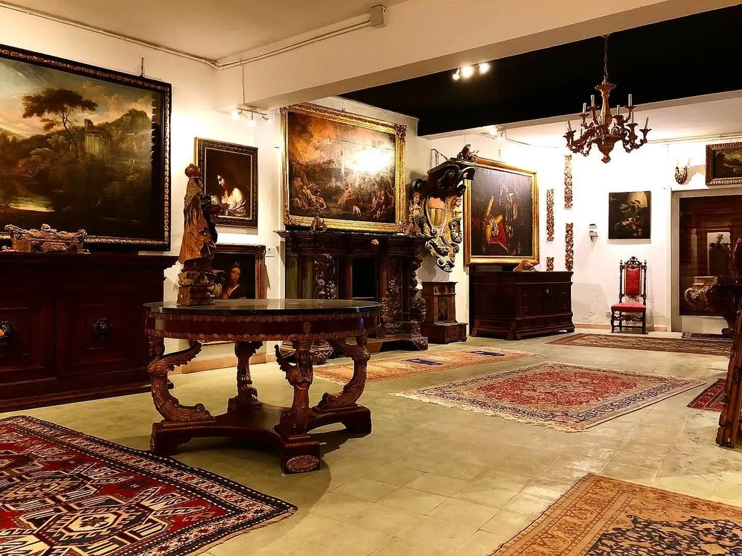 The image is photo-realistic and captures an antique gallery or museum setting with various artifacts. Key elements such as the large central table and prominent paintings are adequately in focus. The composition effectively showcases a range of items within the space. However, the lighting creates some reflections on the large central painting, which slightly obscures parts of it. Overall sharpness could be improved, and some areas of the room appear somewhat dim.
