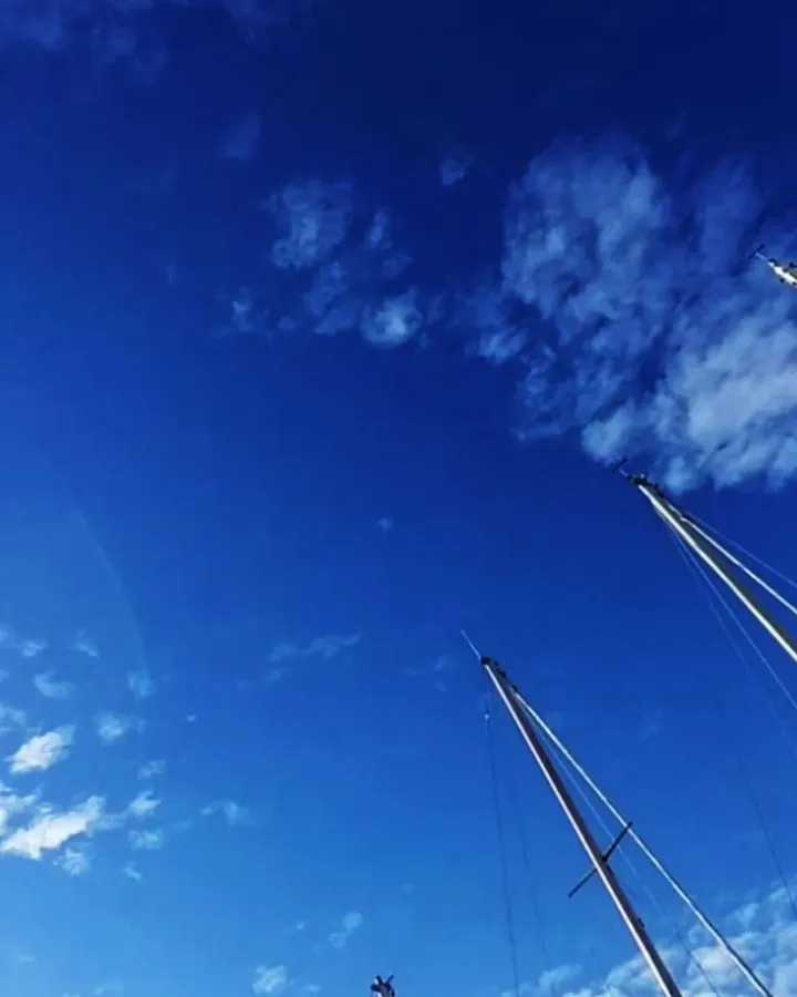 An upward view of a vibrant blue sky with scattered white clouds and the masts of boats.