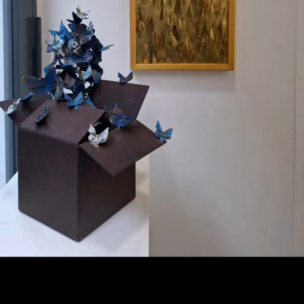 A photograph showcasing two distinct art pieces: a sculpture of blue butterflies emerging from a dark box, and a gold-toned framed artwork depicting abstract, sharp forms, possibly representing grass or spikes. The background is a plain white wall.