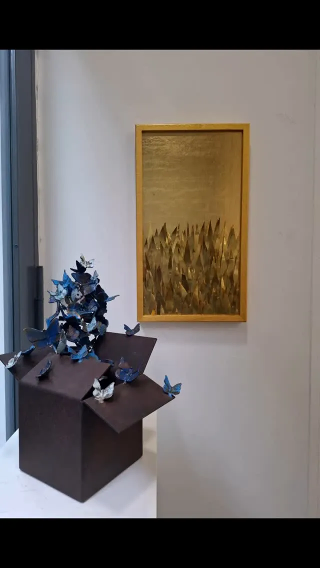 A photograph showcasing two distinct art pieces: a sculpture of blue butterflies emerging from a dark box, and a gold-toned framed artwork depicting abstract, sharp forms, possibly representing grass or spikes. The background is a plain white wall.