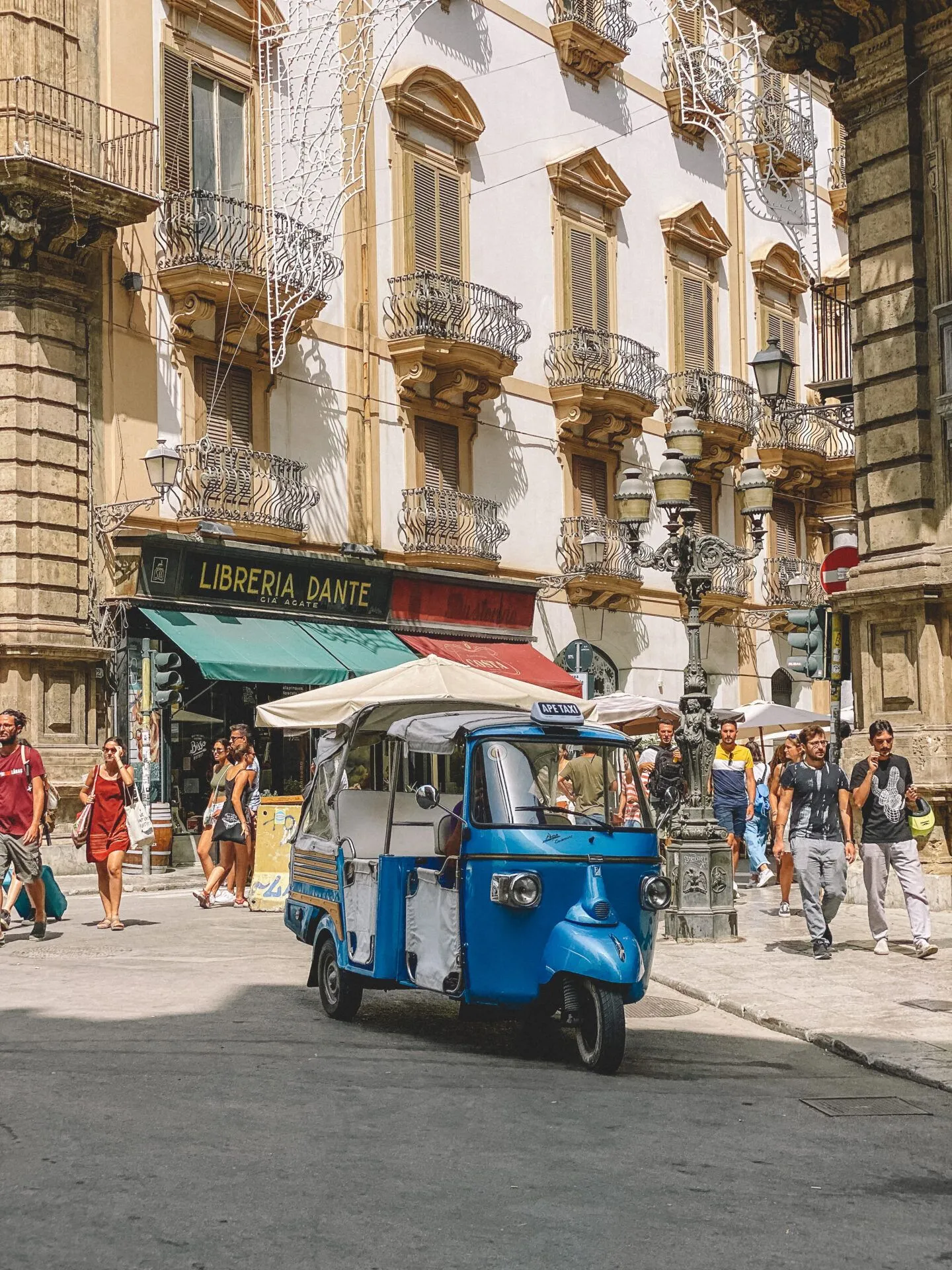 An image depicting a blue tuk-tuk parked on a city street in front of a building with ornate balconies and a bookstore. Several pedestrians are walking in the background.