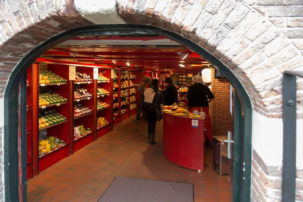 This is a photo-realistic, editorial-style image capturing the interior of a specialty food shop, likely selling cheese, framed through an archway. The composition effectively draws the viewer into the scene, with the key products on shelves and the service counter well in focus and appearing sharp. The colors are accurate, showcasing the vibrant red shelving and authentic product hues. The primary area of improvement lies in the lighting, which is somewhat uneven; the foreground is slightly dark, and there are a few bright spots that could be better managed. Overall, it's a good quality image depicting an allowed subject matter without any unwanted elements.