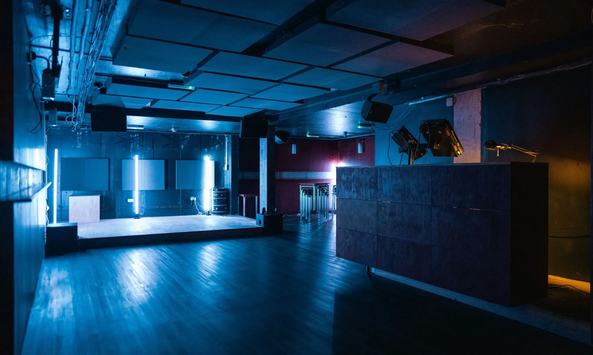 An empty club or venue interior, illuminated by blue ambient lighting, showcasing a stage area with vertical light fixtures and a counter. The image has a moody atmosphere.