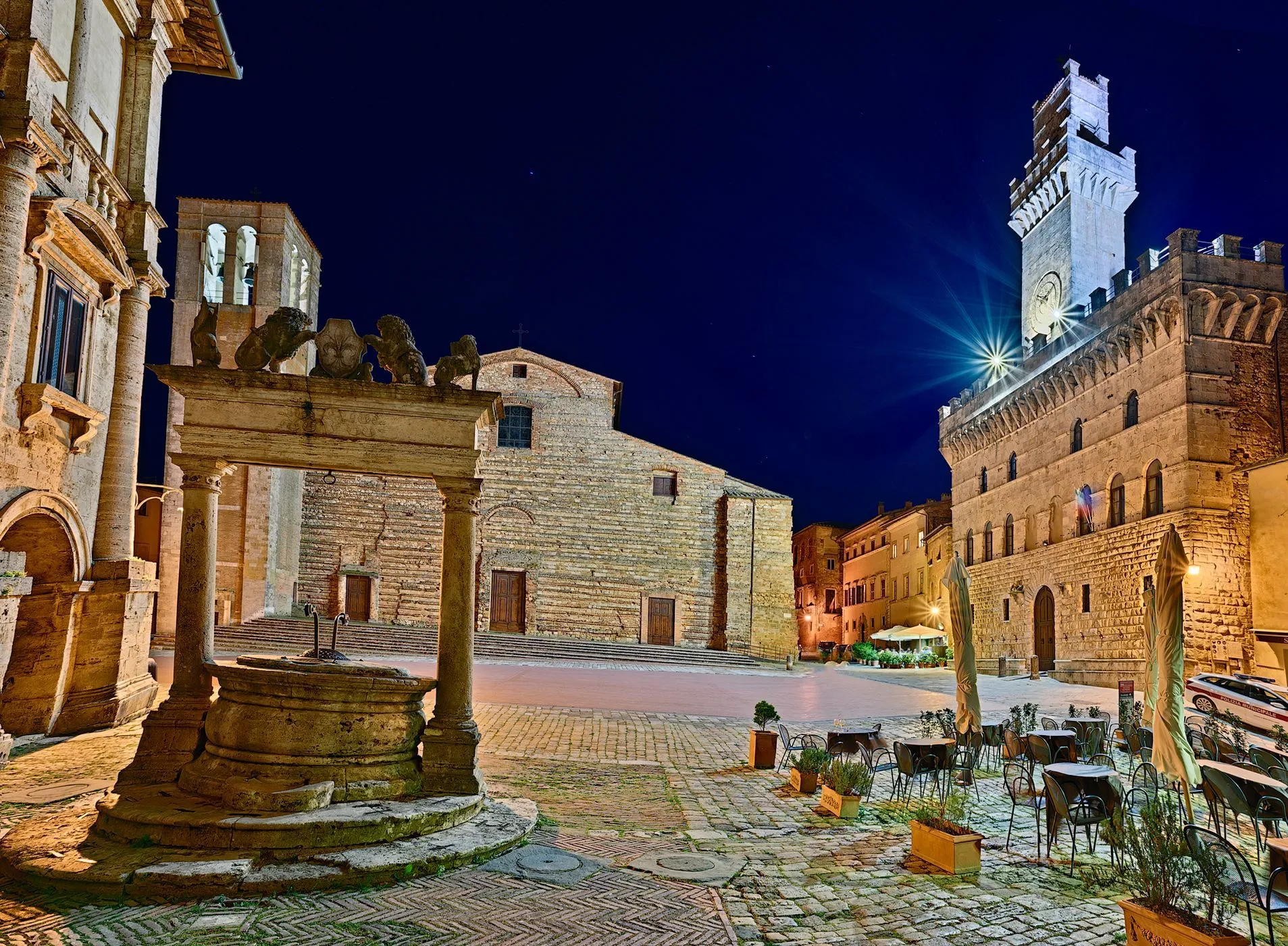 An atmospheric night shot of a historic European town square, featuring old stone buildings, a decorative well in the foreground, and cobblestone paving. The scene is illuminated by artificial lights, creating a warm glow.