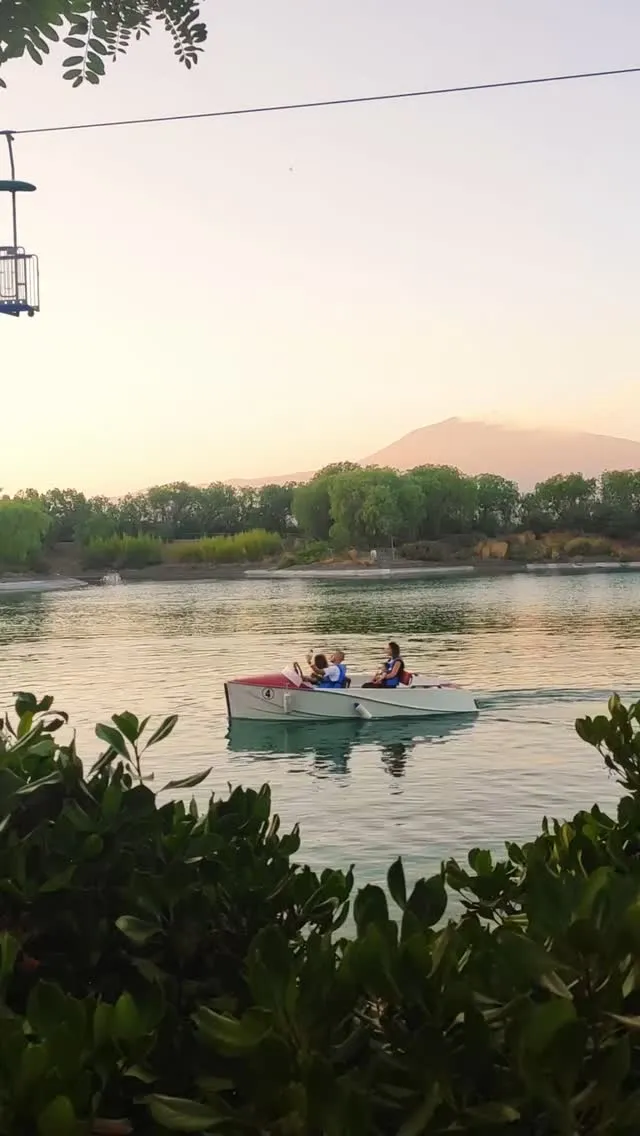 The image captures a peaceful scene with a boat carrying people on a calm lake, set against a backdrop of trees and a distant mountain under soft, warm lighting. The overall mood is serene and natural.