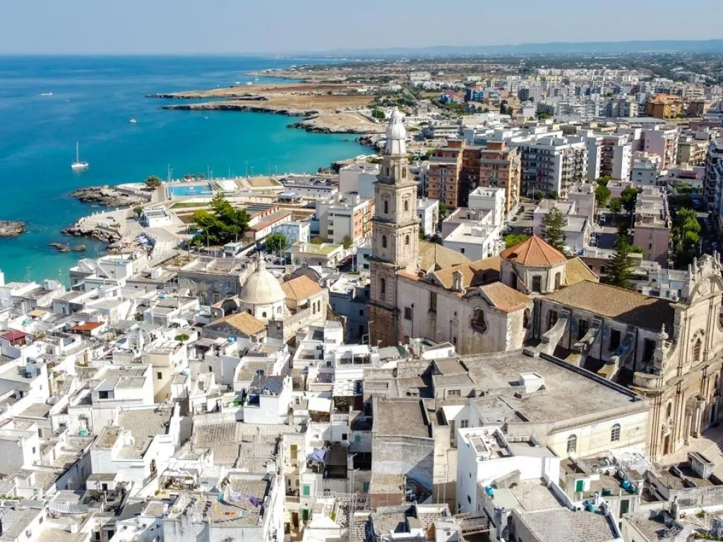 An aerial view of a coastal Mediterranean town with white buildings, a prominent church tower, and a vibrant blue sea.