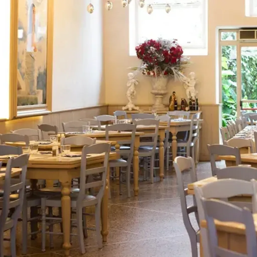 An image capturing the interior of a restaurant with multiple wooden tables and light grey chairs arranged neatly. A large vase with red flowers and decorative statues is visible against a wall. Large windows and a glass door at the back provide a view of lush greenery outside.