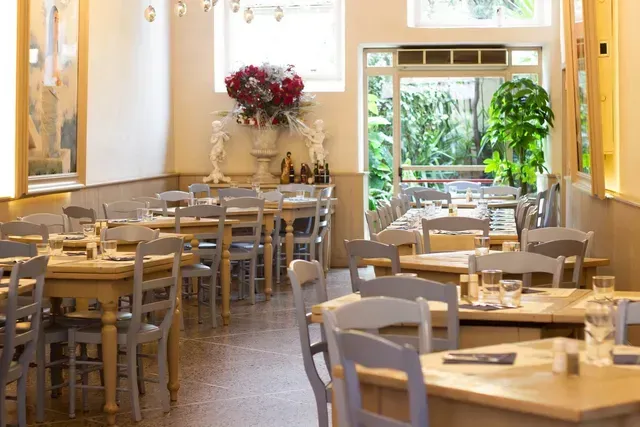 An image capturing the interior of a restaurant with multiple wooden tables and light grey chairs arranged neatly. A large vase with red flowers and decorative statues is visible against a wall. Large windows and a glass door at the back provide a view of lush greenery outside.