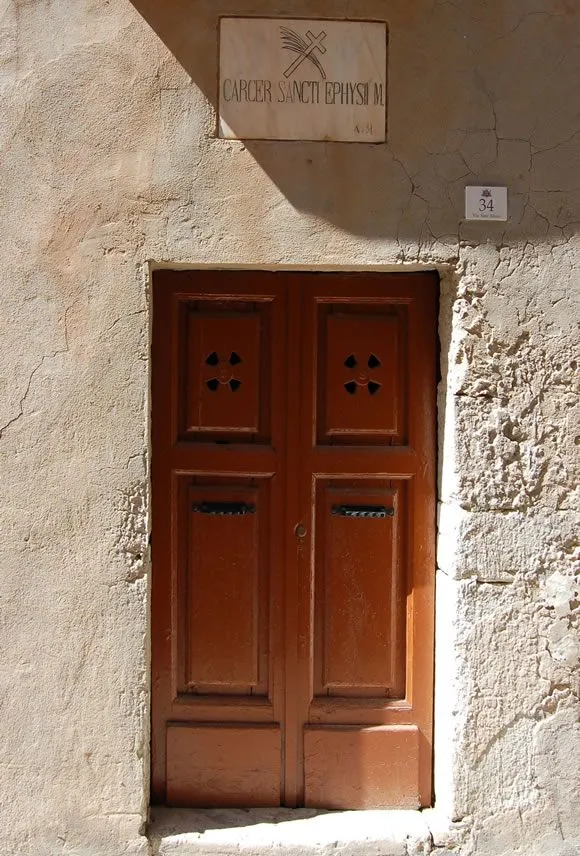 A straight-on, close-up view of an old, brown wooden door with decorative cutouts and mail slots, set into a textured, light-colored stucco or stone wall. A house number '34' is visible above the door on the right side.