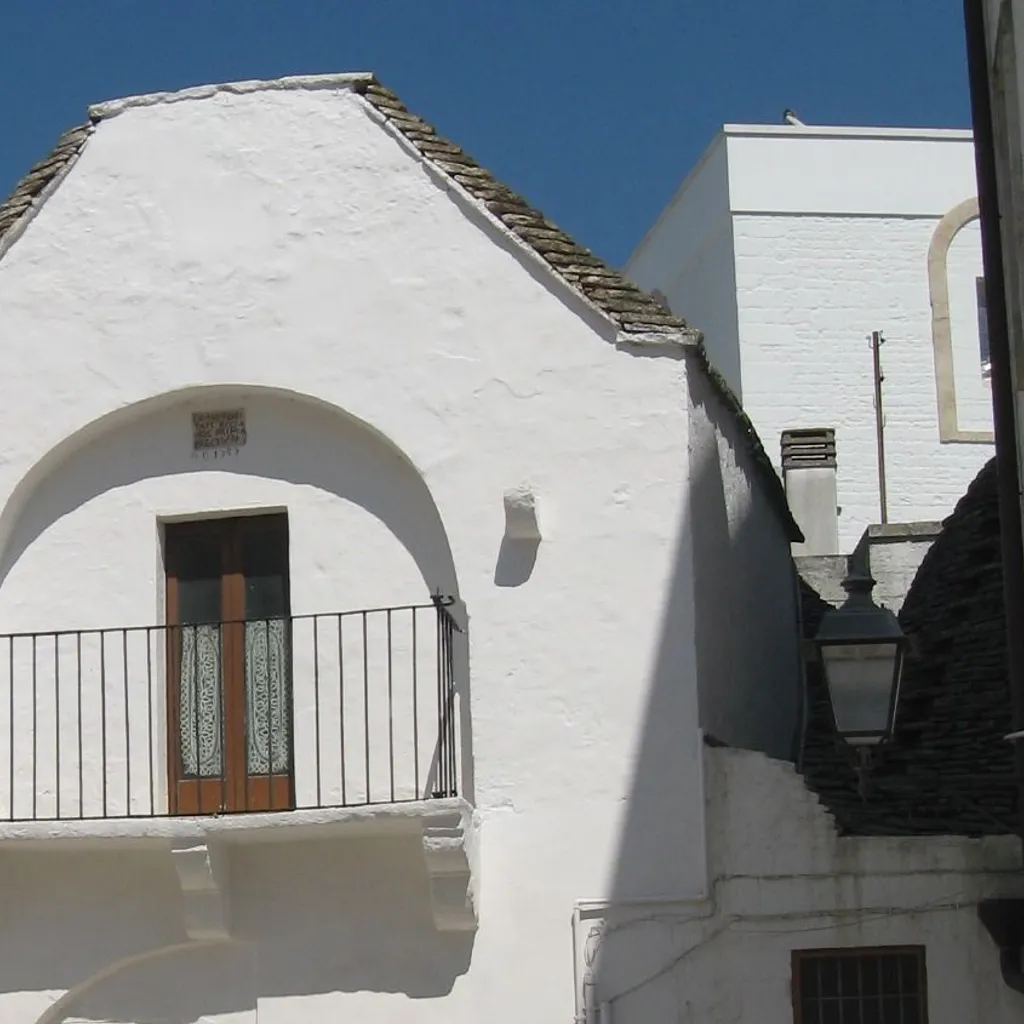 The image showcases a distinctive white building with a balcony and an arched entrance, set against a clear blue sky. The traditional architecture is well-captured, highlighting the building's unique features.
