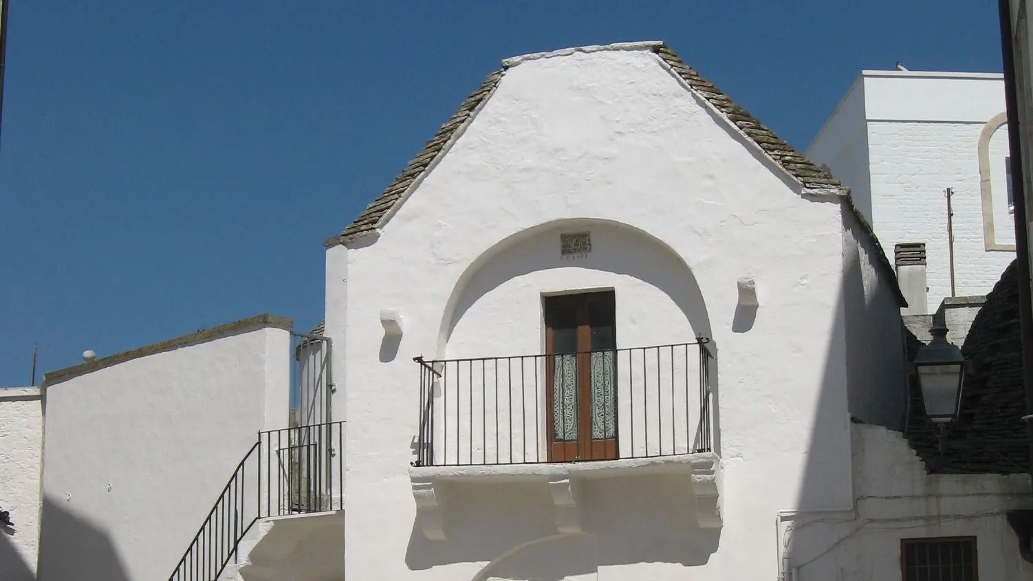 The image showcases a distinctive white building with a balcony and an arched entrance, set against a clear blue sky. The traditional architecture is well-captured, highlighting the building's unique features.