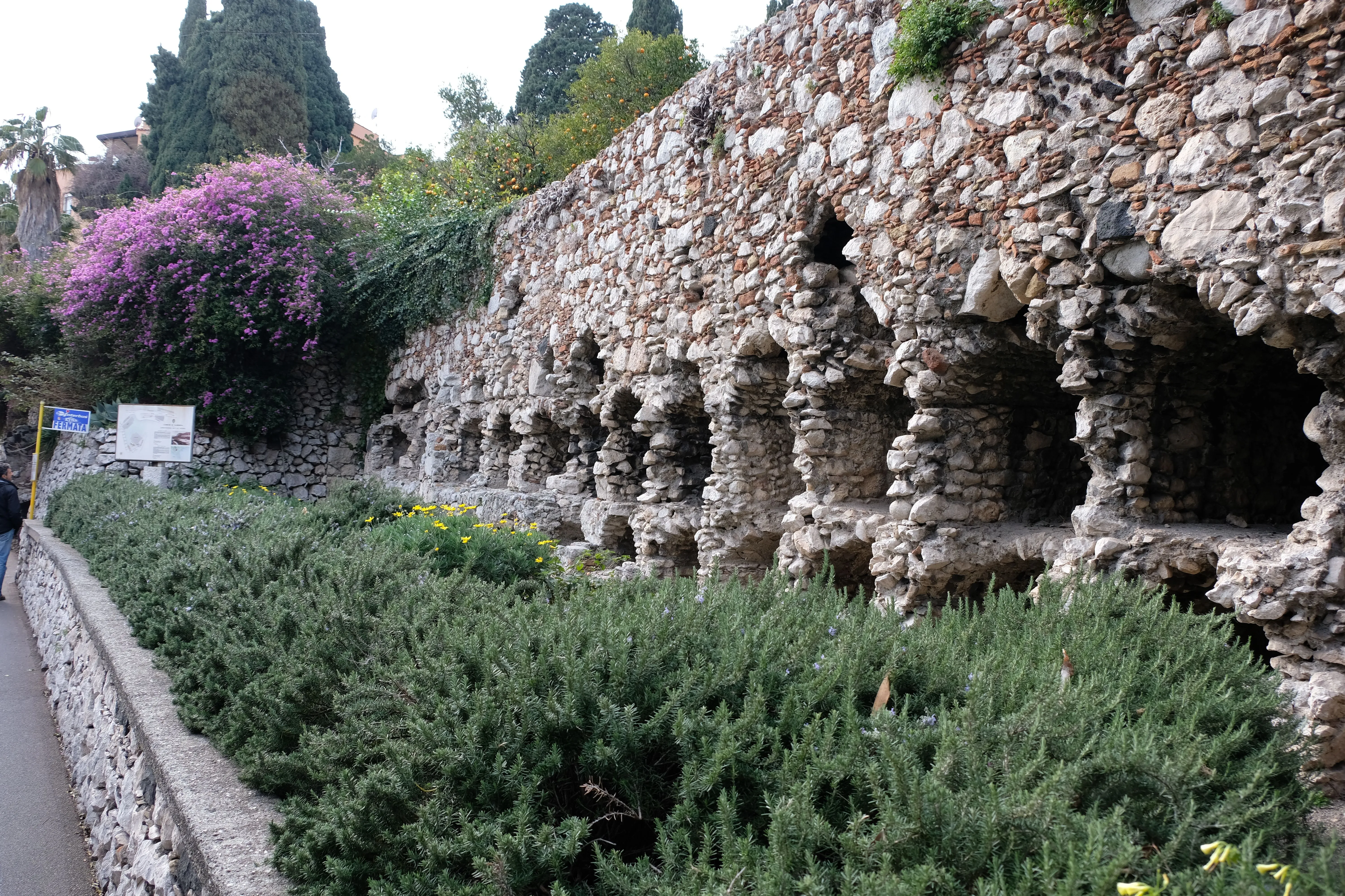 An editorial-style image showcasing an ancient stone wall with arched alcoves, possibly ruins of a historical structure, surrounded by lush green foliage and trees in a garden setting.