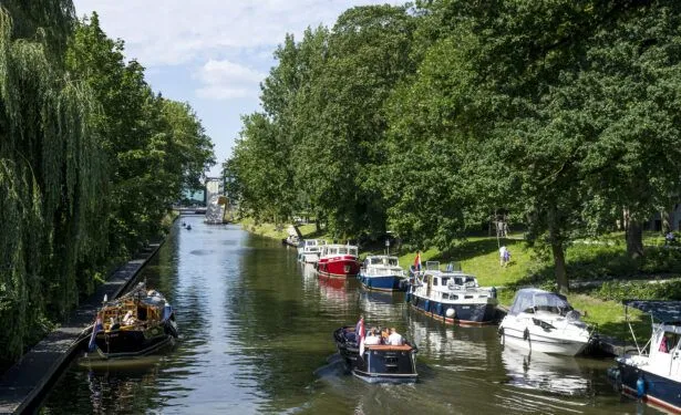 An inviting view of a canal lined with lush green trees and several boats, creating a serene and picturesque outdoor scene. The water reflects the vibrant greenery and clear skies.