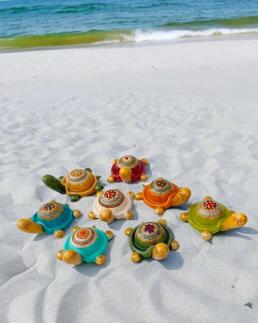 A collection of colorful decorative turtle figurines arranged on white sand with the ocean and waves in the background.