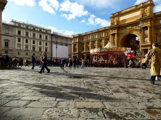 The image effectively captures a bustling European piazza under natural light, showcasing good color accuracy and photo-realism. The historic buildings and the carousel are sharply in focus, creating a clear sense of place. However, the composition is slightly weakened by a prominent, empty white screen on the left side, which acts as a distracting element. Additionally, some moving individuals show motion blur, and the expansive cobblestone foreground, though detailed, occupies a large portion of the frame.