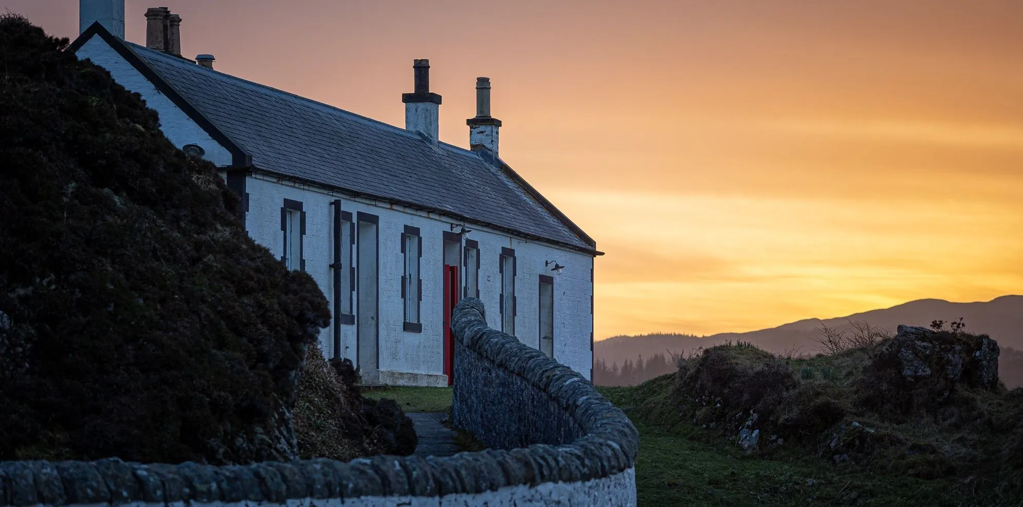 An atmospheric outdoor shot depicting a white, rural house with a distinctive red door and two chimneys, viewed from an angled perspective. A curving stone wall occupies the foreground, leading the viewer's eye towards the house. The sky is filled with warm orange and yellow hues, suggesting either a sunset or sunrise, with distant, misty hills visible on the horizon.