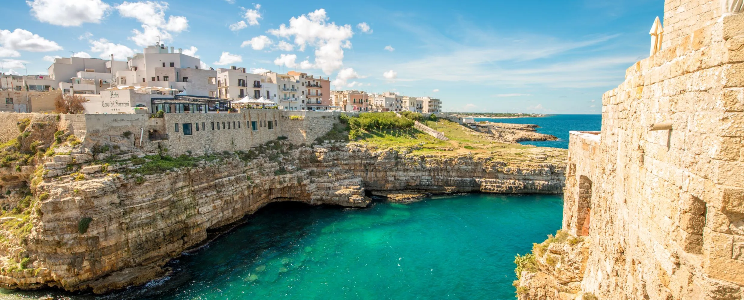 A beautiful panoramic view of a coastal town built atop rugged cliffs, overlooking a stunning turquoise sea under a bright blue sky with scattered white clouds. The ancient buildings with their white and pastel facades contrast beautifully with the vibrant water.